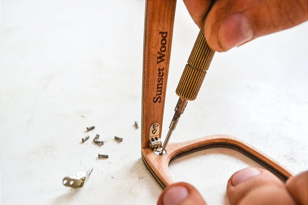 Mãos fixando a dobradiça de uma haste de madeira avermelhada, com o nome Sunset Wood gravado.